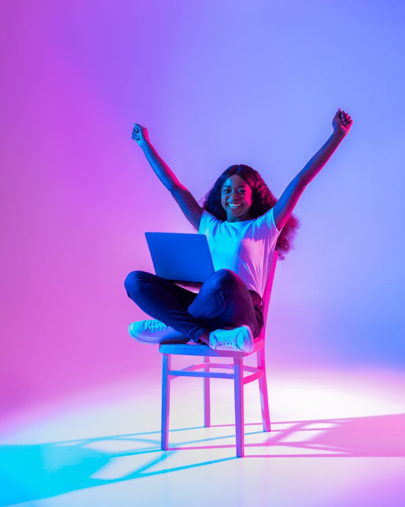 Excited young lady sitting on chair with laptop, raising arms up, celebrating success.